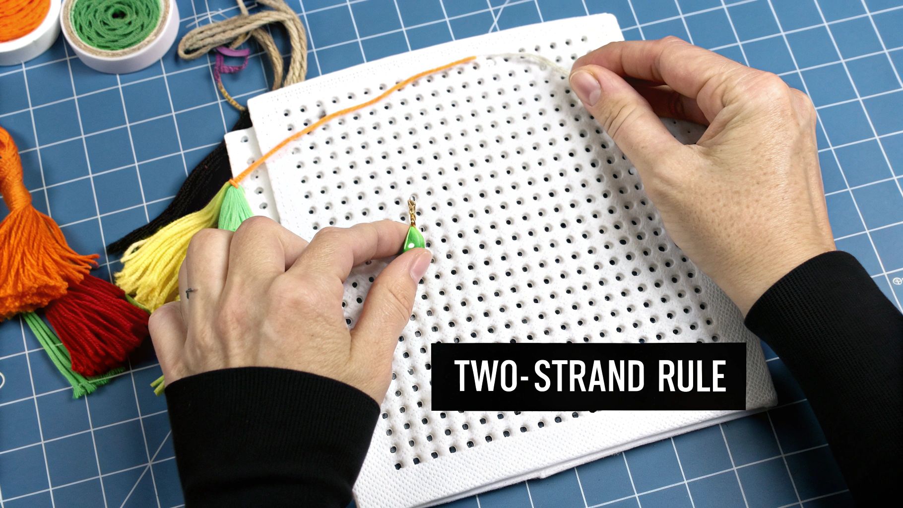 Hands work on a white perforated board with orange thread and a green charm, surrounded by craft supplies.