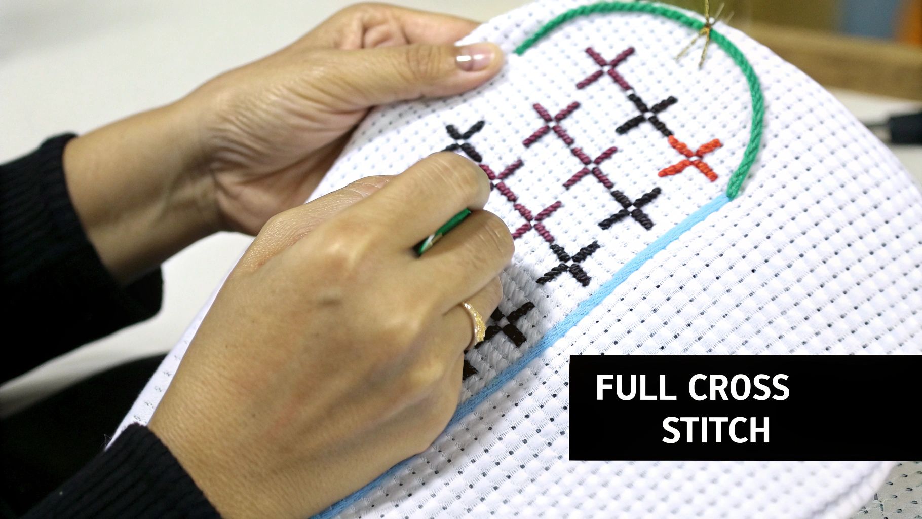 Close-up of hands embroidering full cross stitches in black, purple, and orange on white fabric.