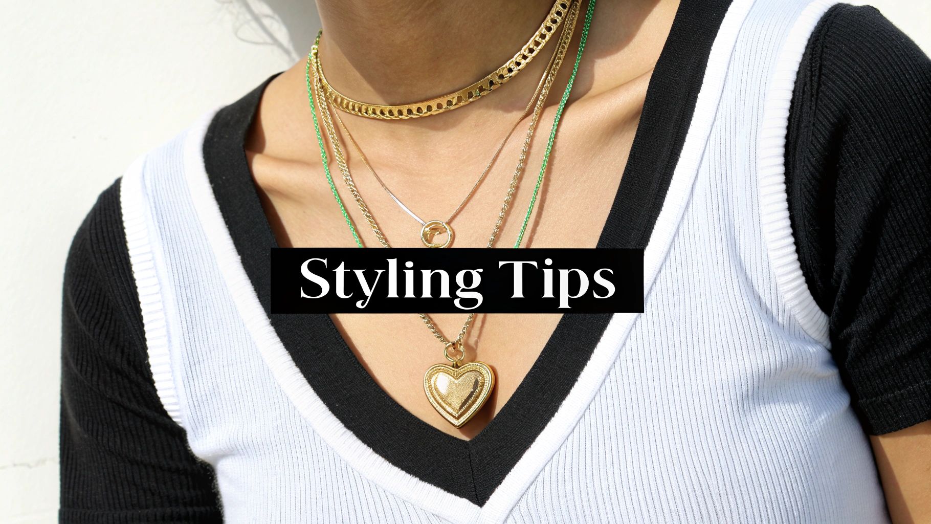 A person wears multiple layered gold necklaces, including a heart-shaped locket, over a ribbed v-neck top.