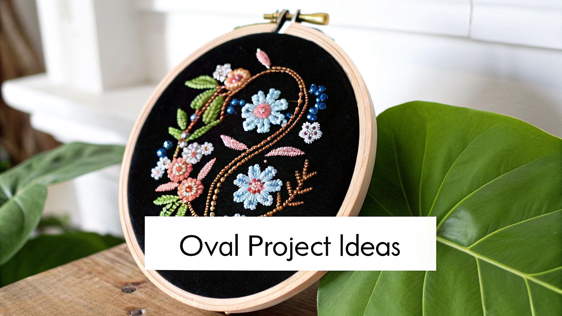 An oval embroidery hoop with colorful floral beadwork and stitching, displayed on wood with a plant.