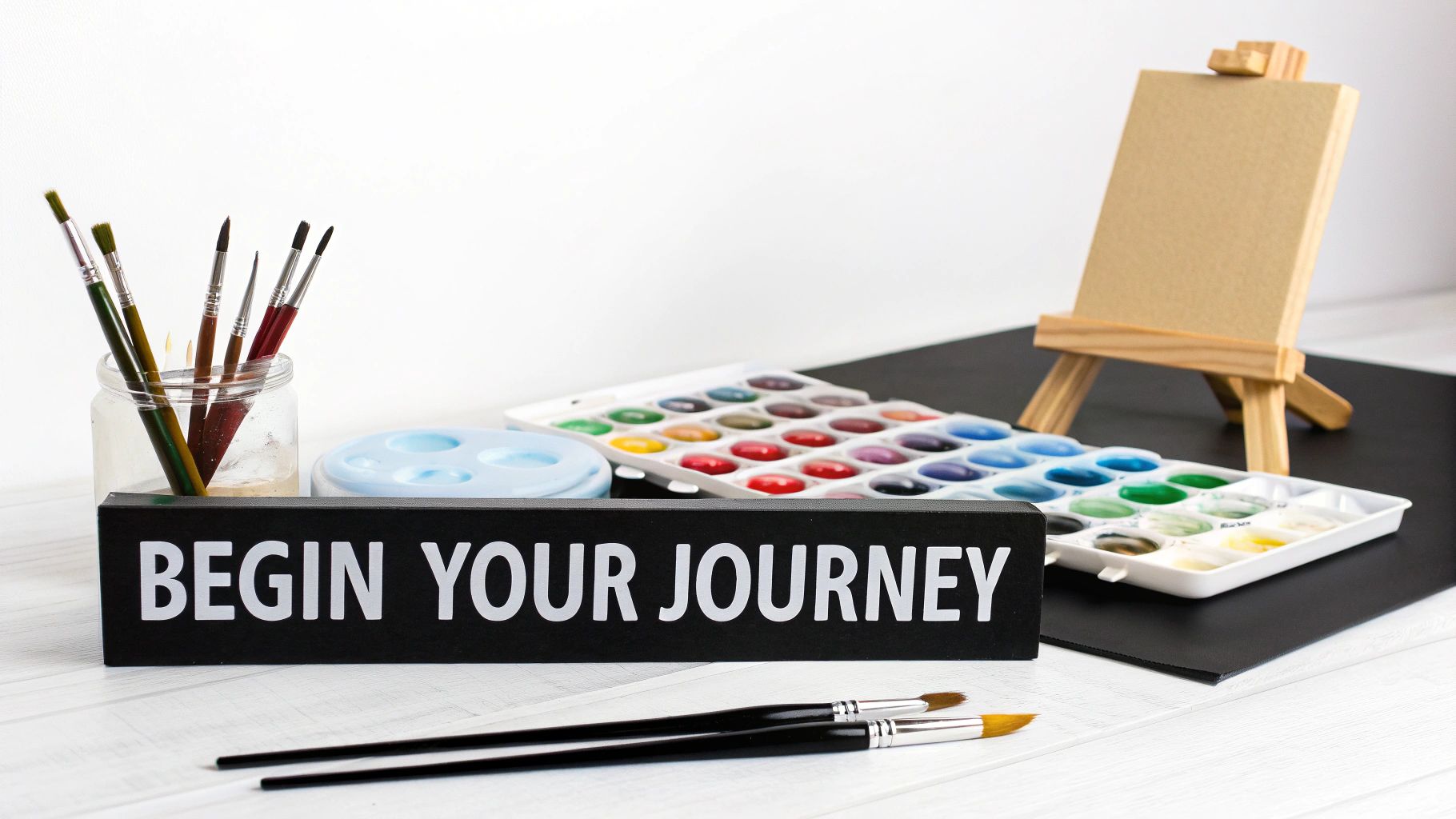 A table setup with watercolor paint sets, paintbrushes in a jar, a mini easel, and a sign that reads 'BEGIN YOUR JOURNEY'.