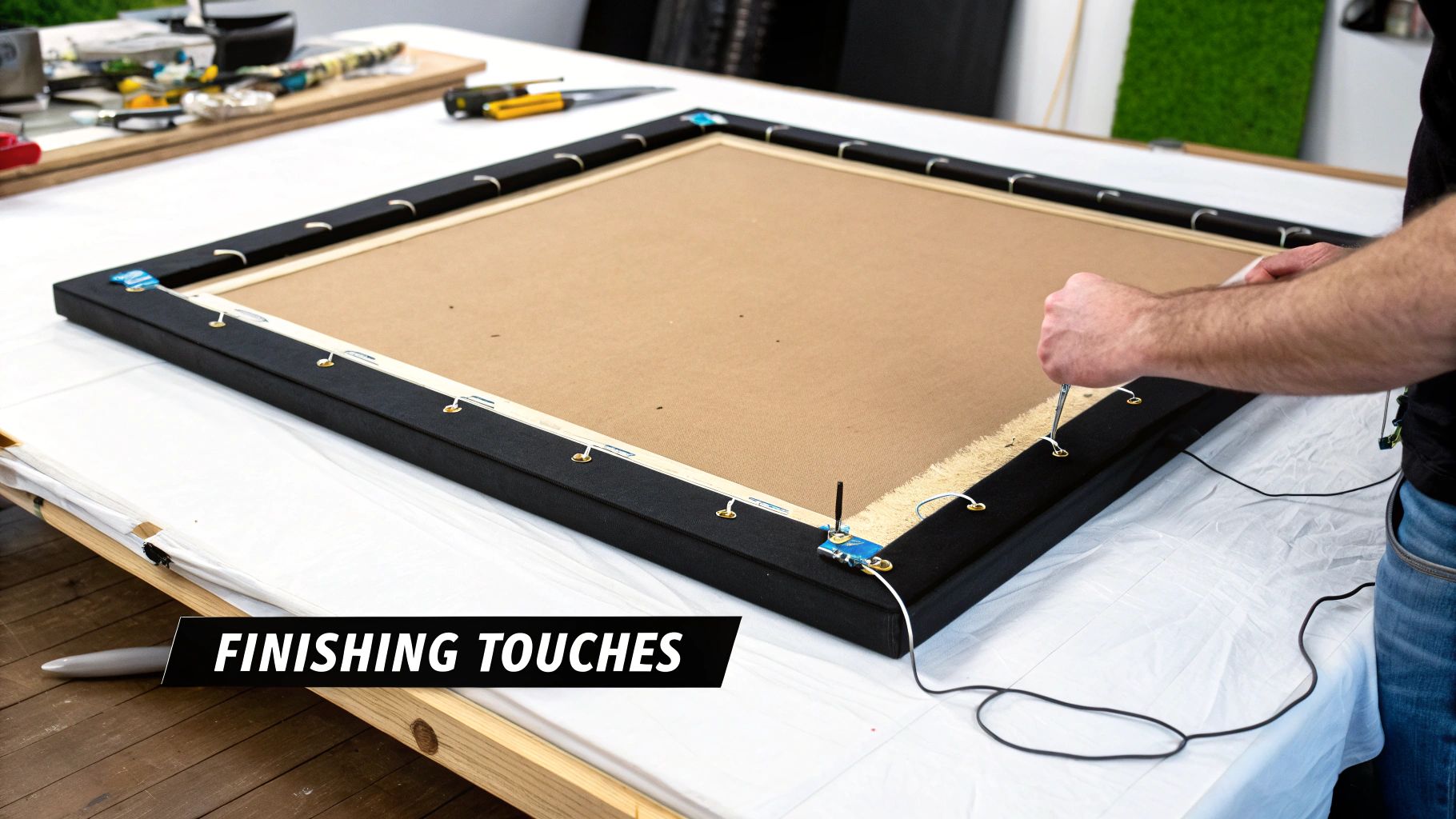 A person is attaching hanging hooks and wire to the back of a framed canvas on a white table.