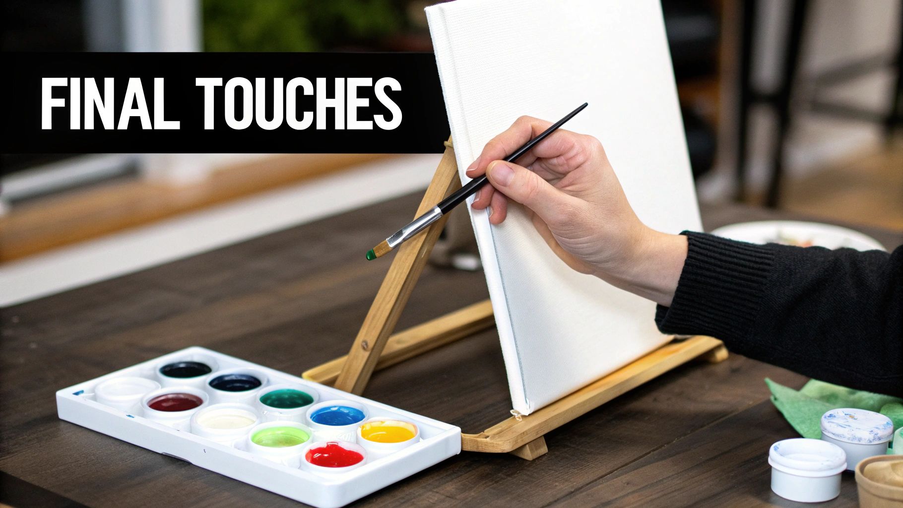 A hand holds a paintbrush with green paint over a blank canvas, next to a colorful paint palette.