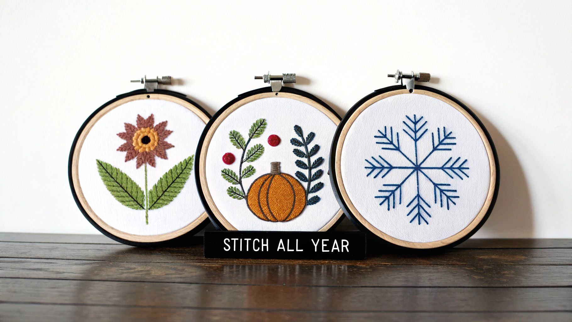 Three embroidery hoops with seasonal cross-stitch designs: a flower, pumpkin, and snowflake, with 'STITCH ALL YEAR' text.