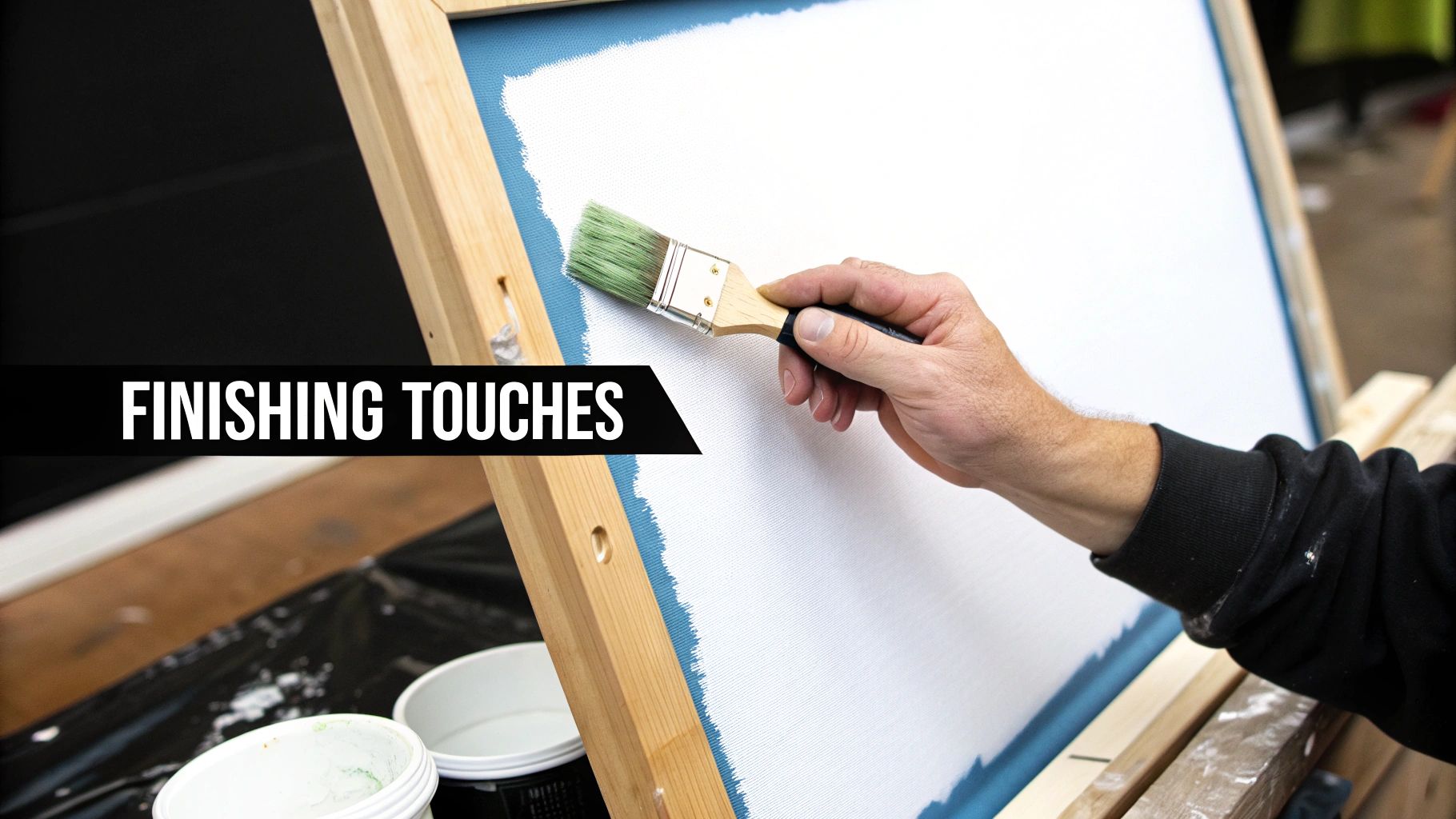 A person's hand holding a green paintbrush, adding finishing touches to a white canvas on an easel.