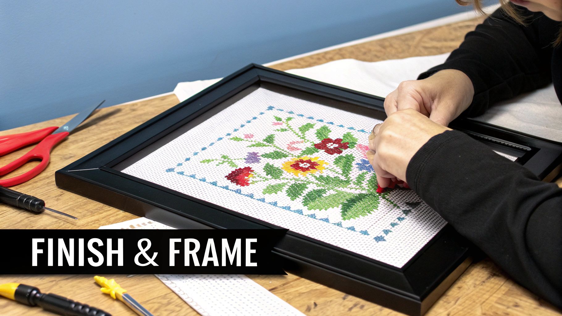 A person's hands framing a colorful floral cross-stitch project on a wooden table with crafting tools.
