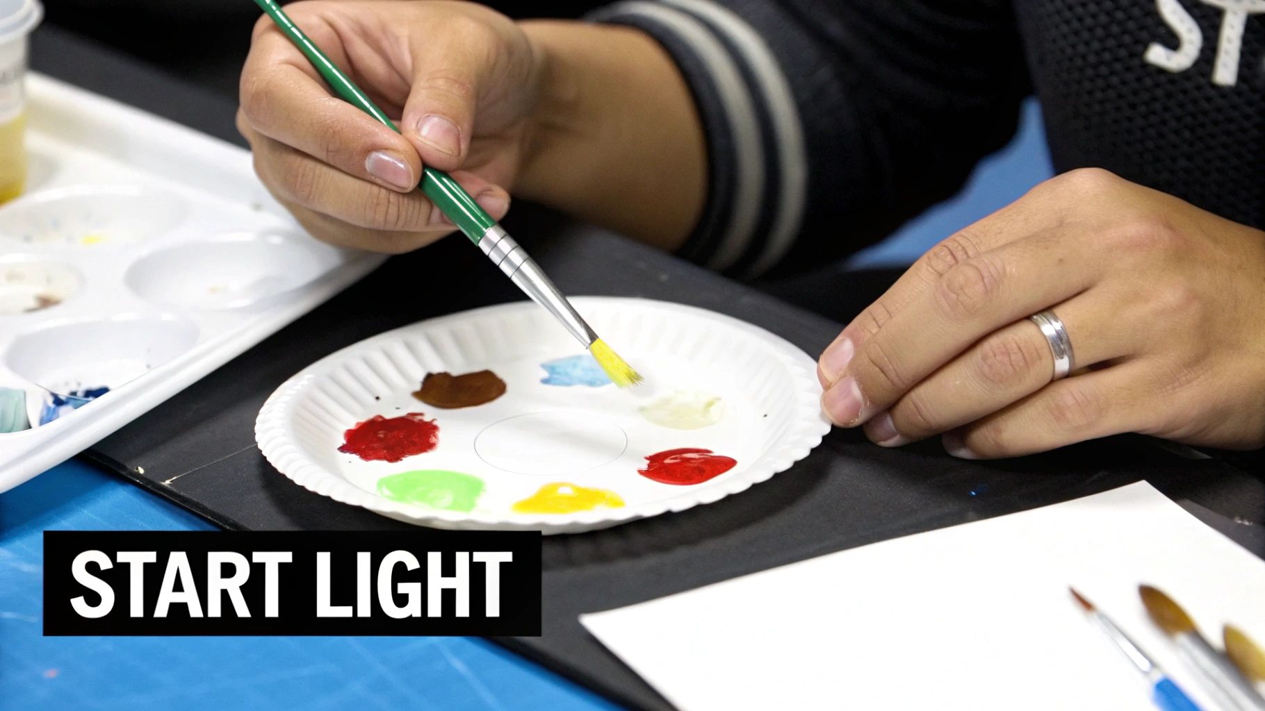 A person's hand holds a green paintbrush, mixing yellow paint on a paper plate palette with other colors.