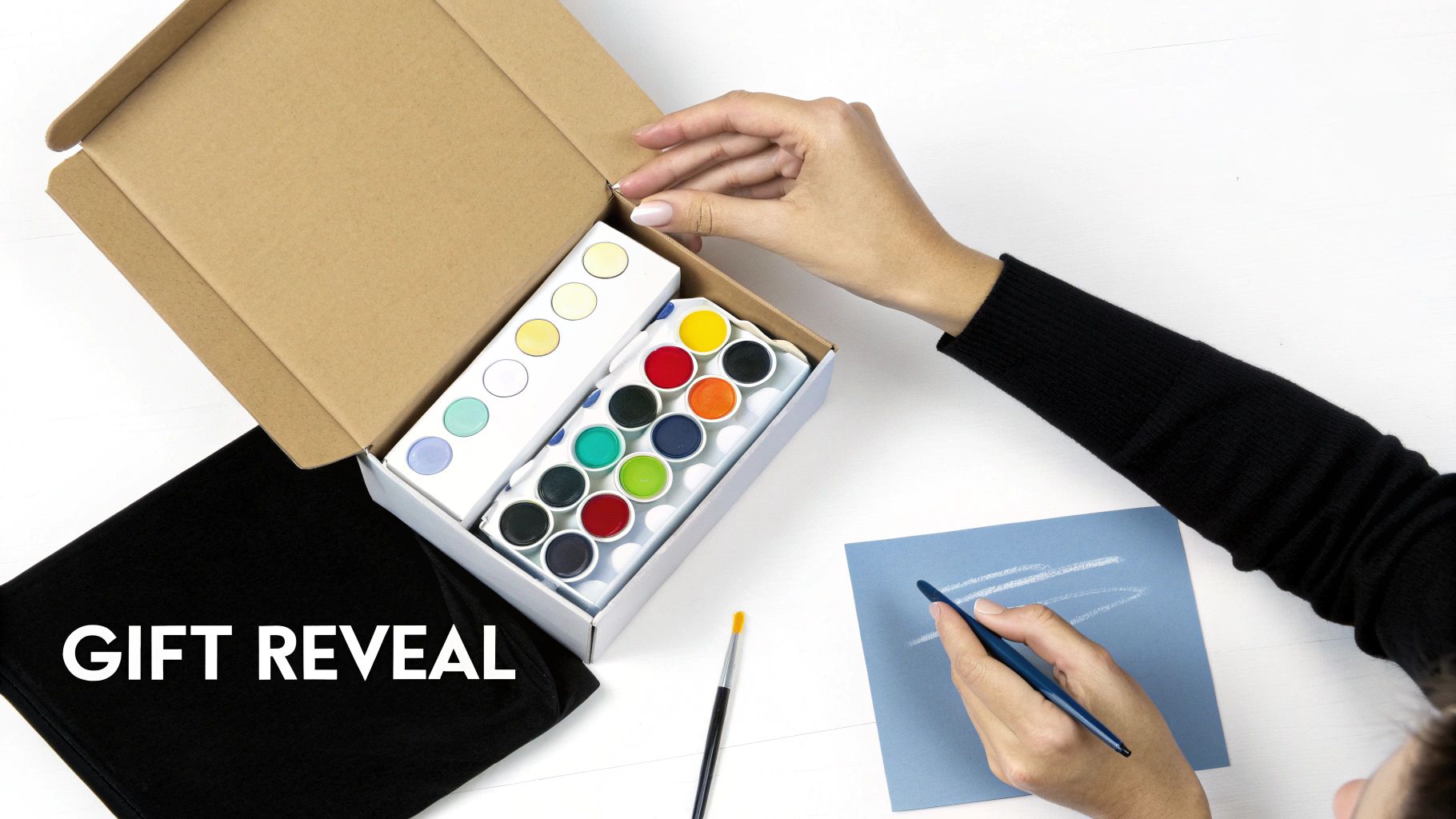 A person's hands open a gift box revealing colorful watercolor paint sets, with another hand drawing.