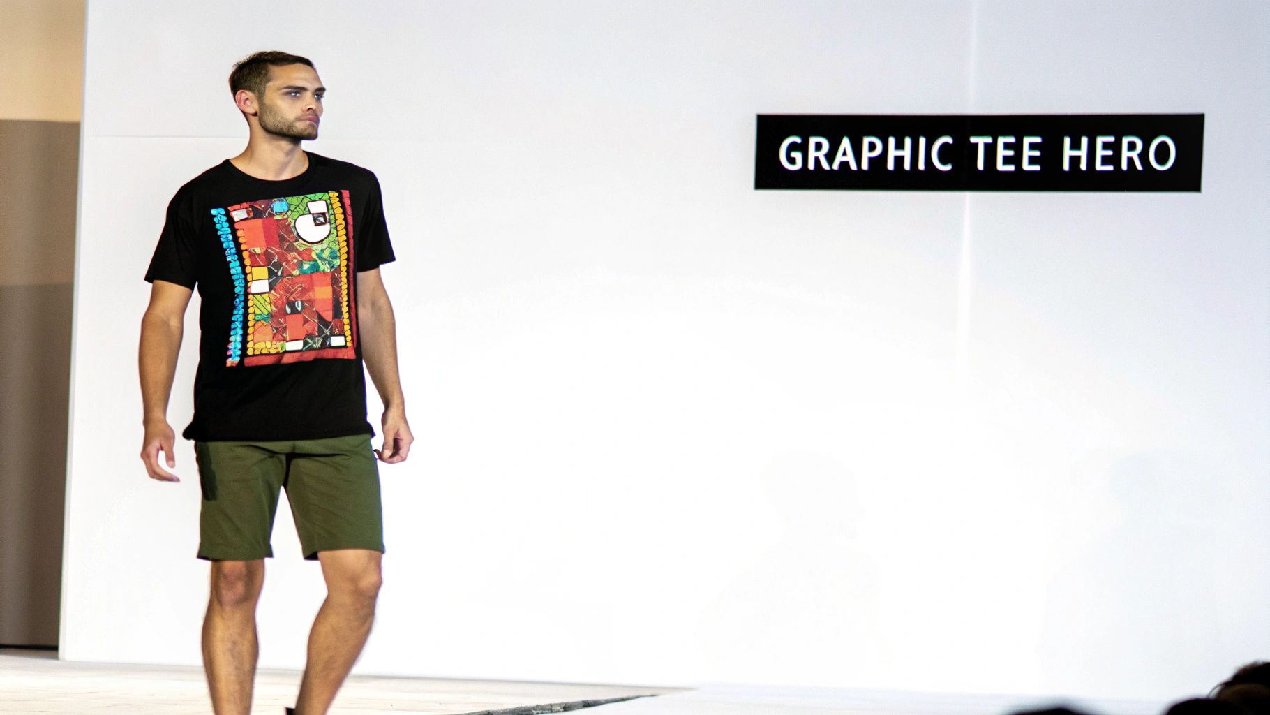 A male model on a runway, wearing a black graphic t-shirt and olive green shorts.