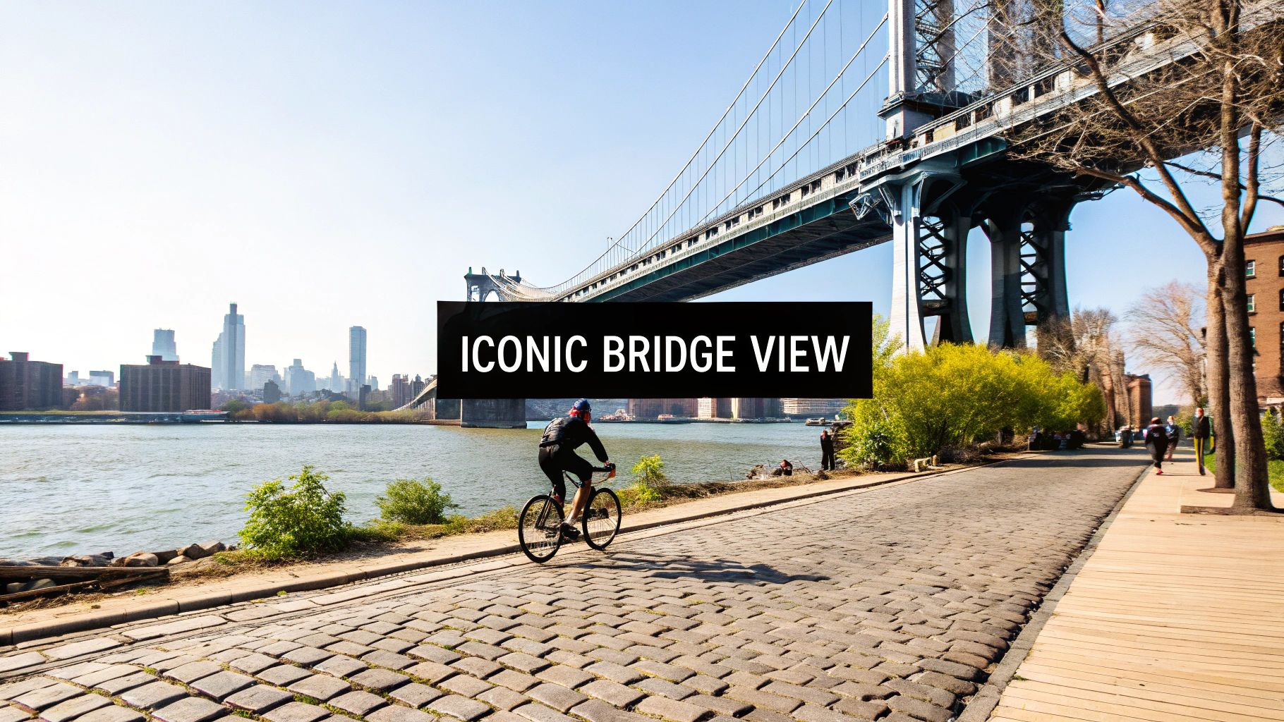 A person cycles on a cobblestone street along a river with an iconic bridge and city skyline in the background.