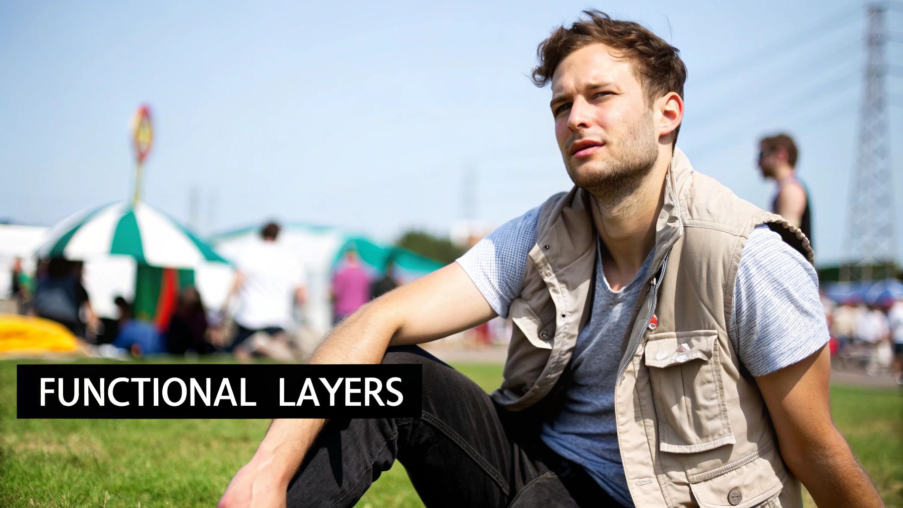 A young man in a beige vest and grey t-shirt sits on grass at an outdoor festival.