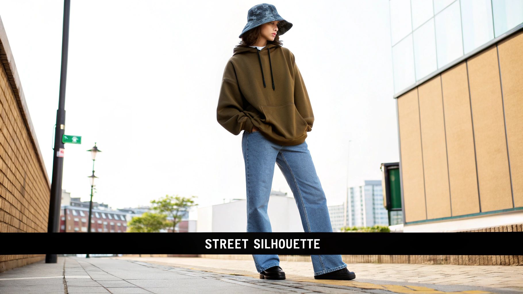 A stylish person in an oversized brown hoodie, light blue jeans, and a bucket hat on a city street.