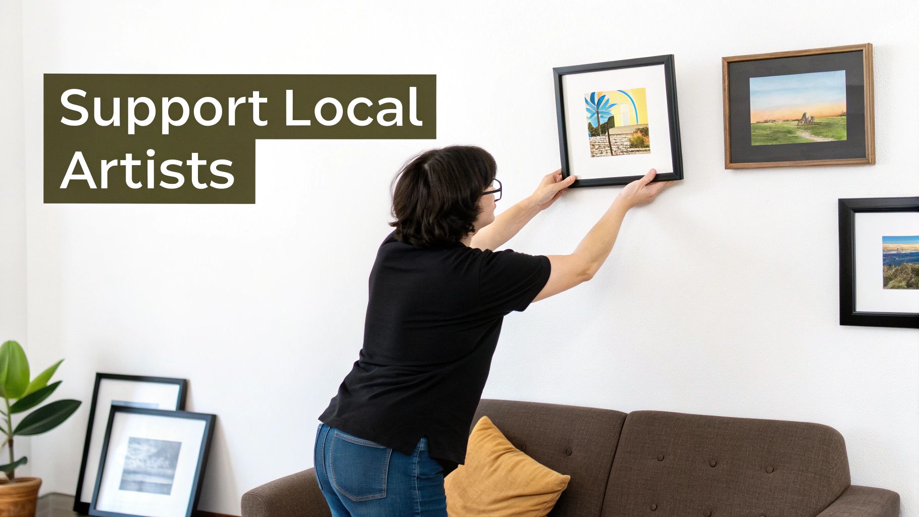 A woman carefully hangs a framed painting on a white wall above a brown sofa, with 'Support Local Artists' text overlay.