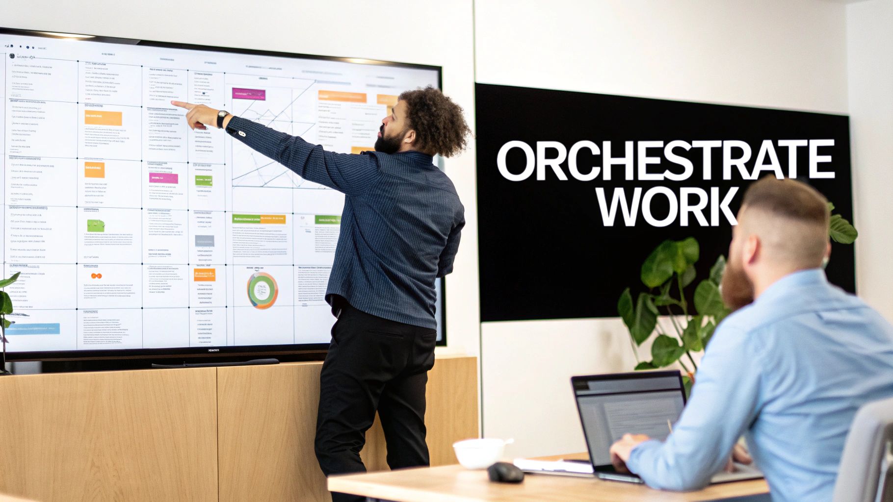 Two men collaborate in an office, one pointing at a large digital display while another works on a laptop.