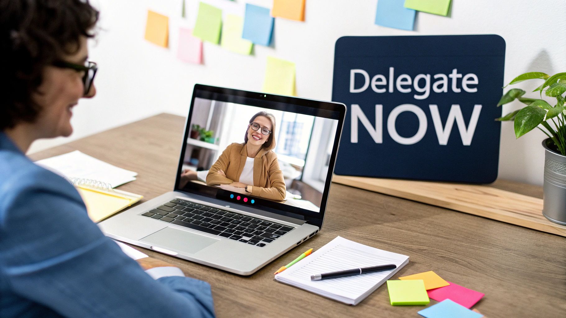 A person smiles during a video call on a laptop, with a 'Delegate NOW' sign behind.