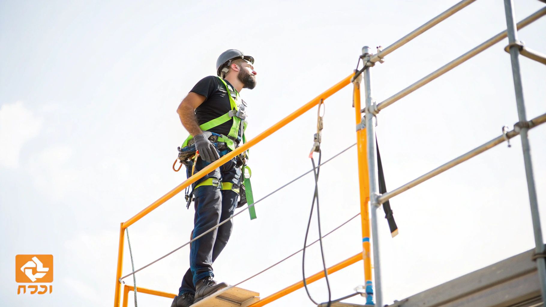 גבר עם קסדה ורתמת בטיחות עומד על פיגומים, מביט מעלה, על רקע שמיים בהירים.