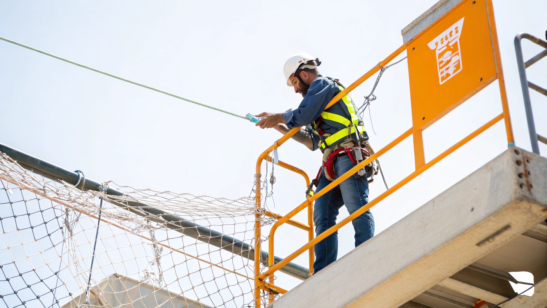 פועל בניין עם קסדה לבנה ורתמת בטיחות צהובה עומד על משטח עבודה גבוה עם חבל.
