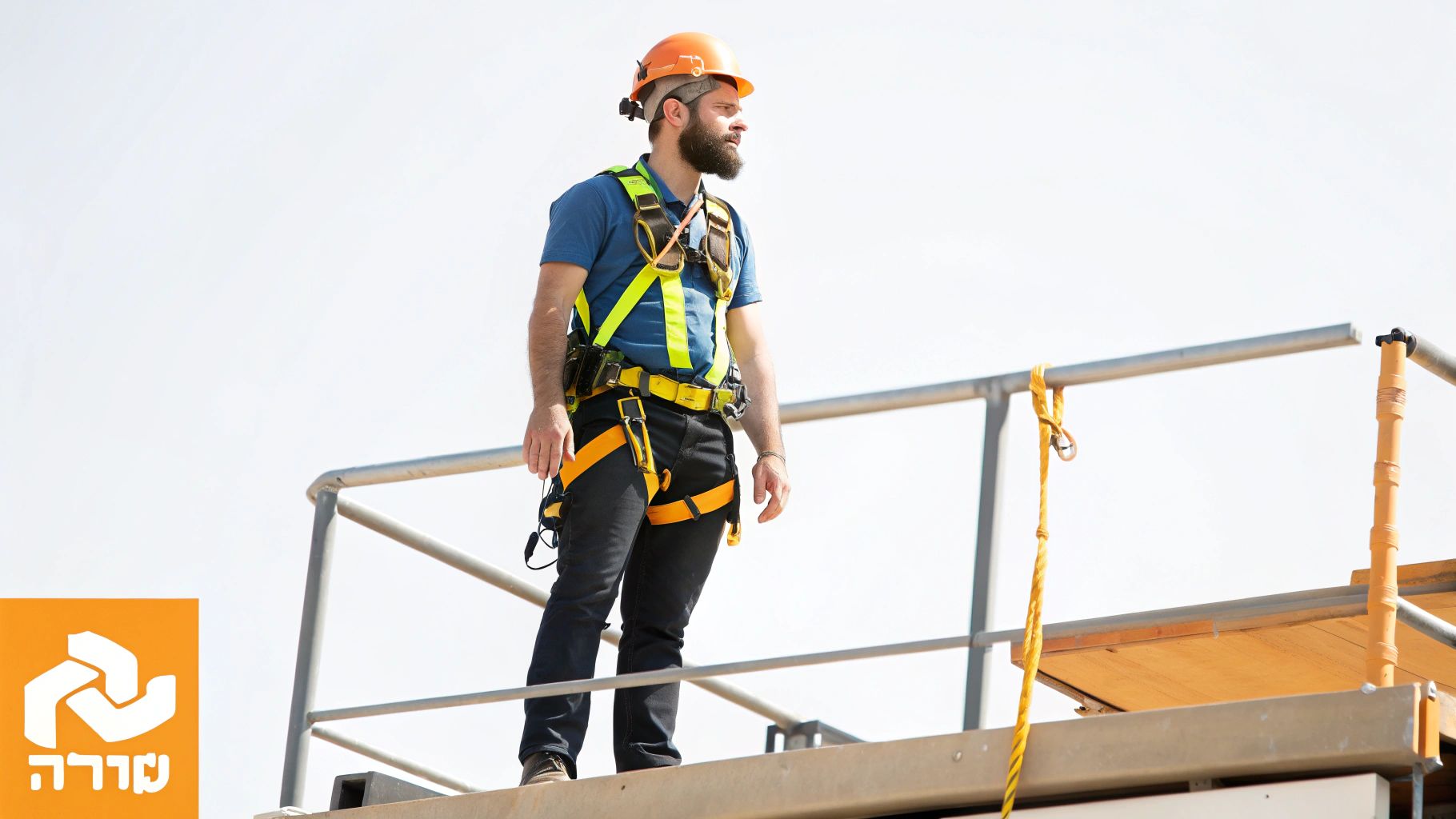 גבר עם זקן, קסדת בטיחות כתומה ורתמת בטיחות צהובה-שחורה עומד על פיגום ומביט הצידה.