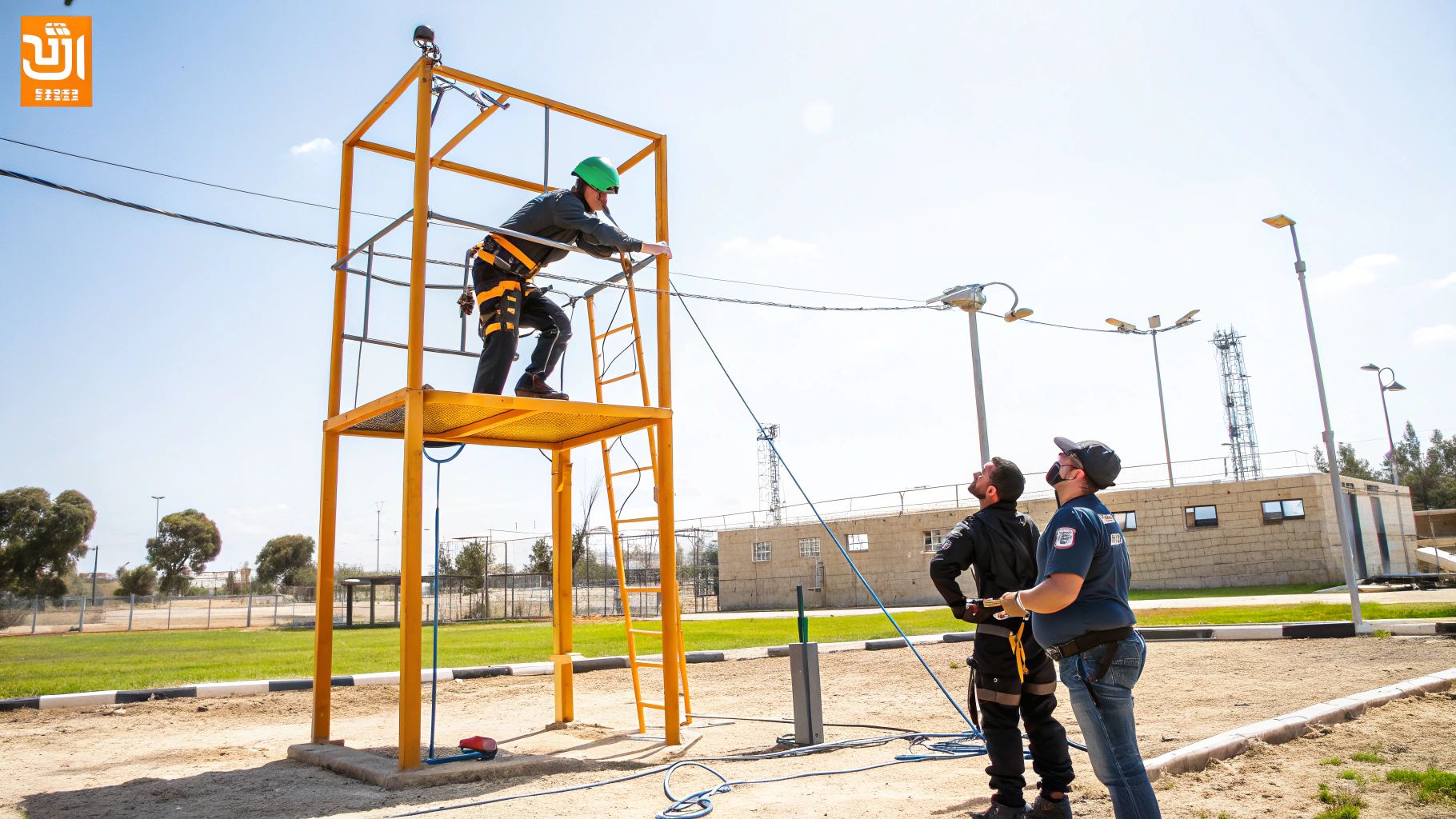 אדם בציוד בטיחות מתאמן בגובה על פלטפורמה, תחת השגחת מדריכים בקורס בטיחות.
